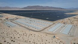 Aerial view of a large solar and battery storage plant set against a dry, arid landscape with mountains and sky visible behind the solar panels and batteries.