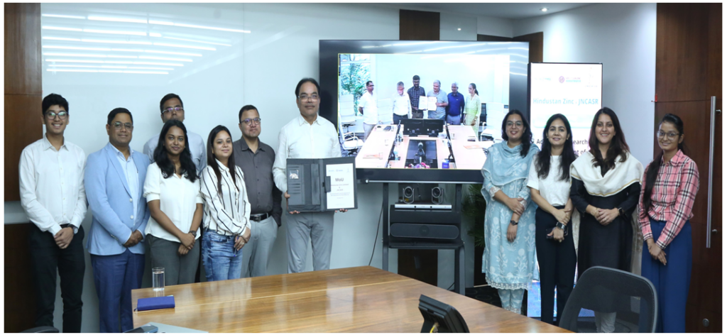 credit indian gov Battery and energy storage researchers standing together in a row in a room with a large screen in the center showing people.